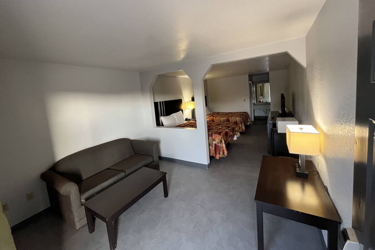 Une chambre d’hôtel avec un canapé, une petite table, une lampe, un lit recouvert d’une courtepointe à motifs, un miroir et un couloir menant à une autre pièce.