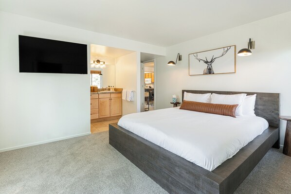 Un dormitorio con una cama grande, un televisor de pantalla plana en la pared, una cocina al fondo y una obra de arte con cuernos de ciervo enmarcada en la pared.