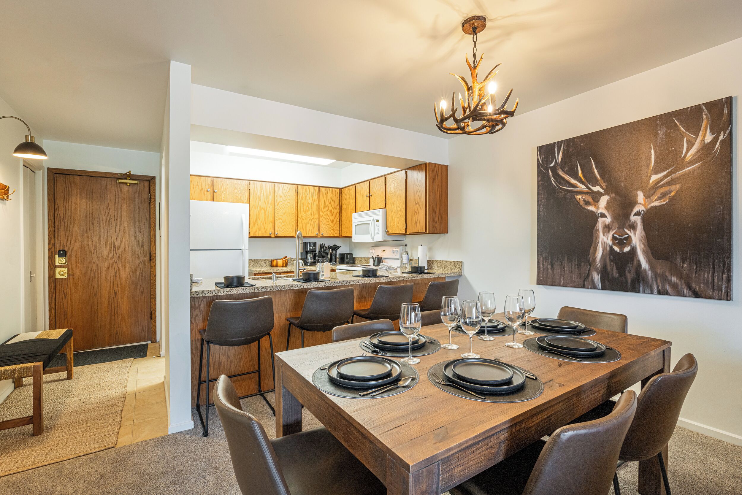 A dining area with a wooden table set for six, a kitchen in the background, and a deer painting on the wall.