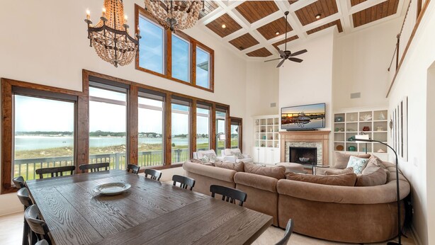 A spacious living room with a large sectional sofa, a dining table with chairs, and a fireplace. There are large windows offering a view of the outdoors. - Hobbs Realty - Very Last Home on the Island w/Heated Pool, Elevator, Luxury Amenities (Holden Beach)