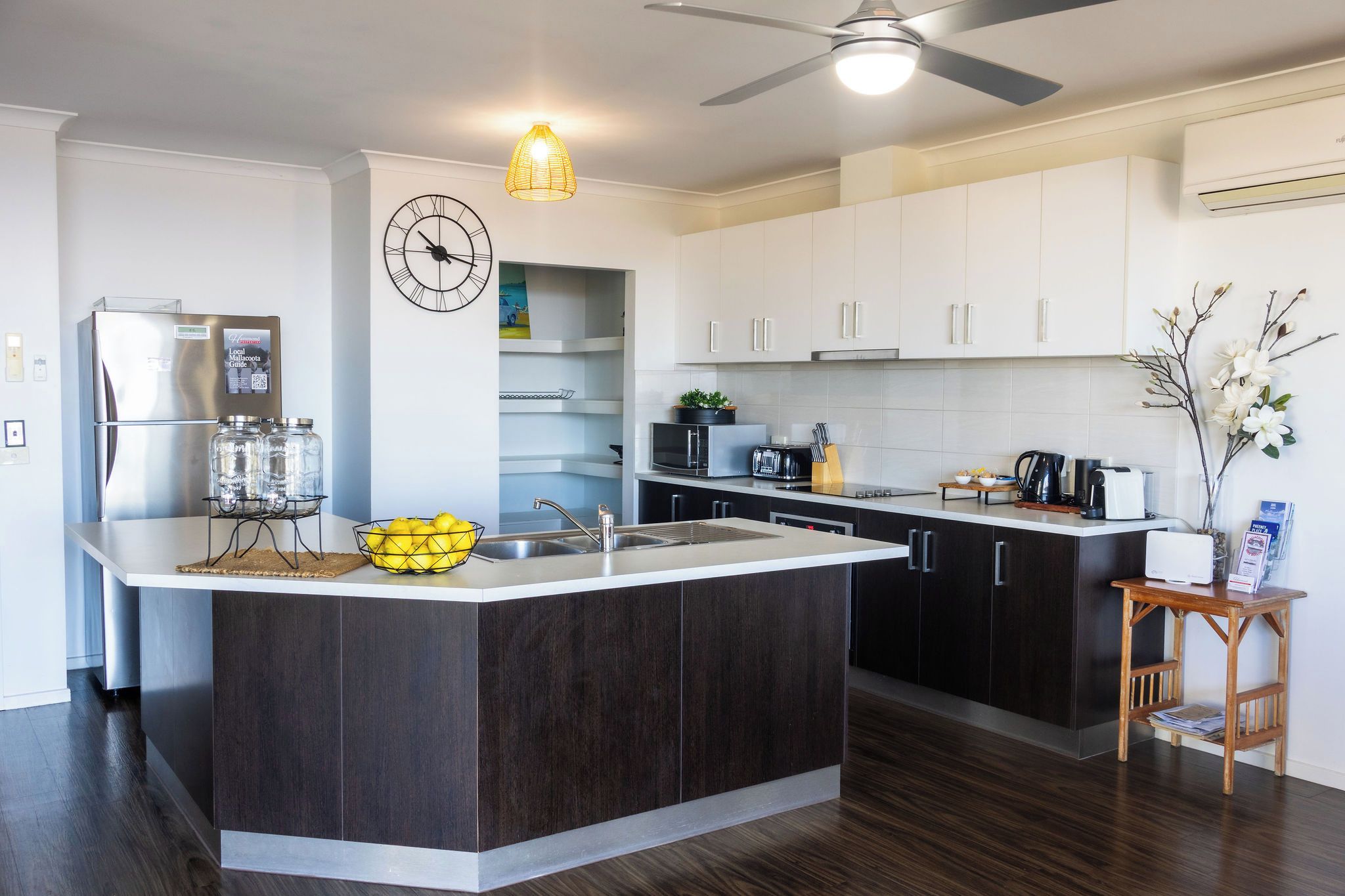 Une cuisine moderne dotée d’un îlot central, de meubles en bois foncé, de plans de travail blancs et d’un ventilateur de plafond.