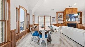 Un coin repas avec une table en bois et des chaises blanches, un canapé blanc et un téléviseur posé sur un meuble en bois.