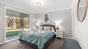 A bedroom with a bed, bedside tables, a bench, a large window with blinds, and a ceiling light fixture. - Kurrajong at Malua (Malua Bay)