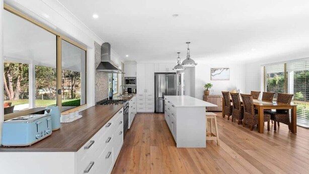 A modern kitchen with a central island, white cabinetry, and a dining area with a table and chairs. - Kurrajong at Malua (Malua Bay)