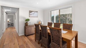 A dining area with a wooden table and woven chairs, a potted plant, and a window with blinds. - Kurrajong at Malua (Malua Bay)