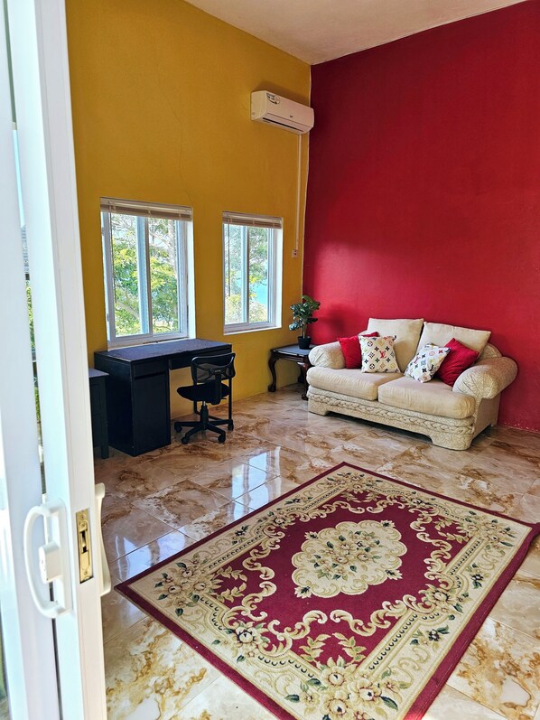 Una stanza con una parete gialla, una parete di colore rosso a contrasto, un divano beige con cuscini decorati e un tappeto ornamentale di grandi dimensioni, oltre a una scrivania con una sedia.