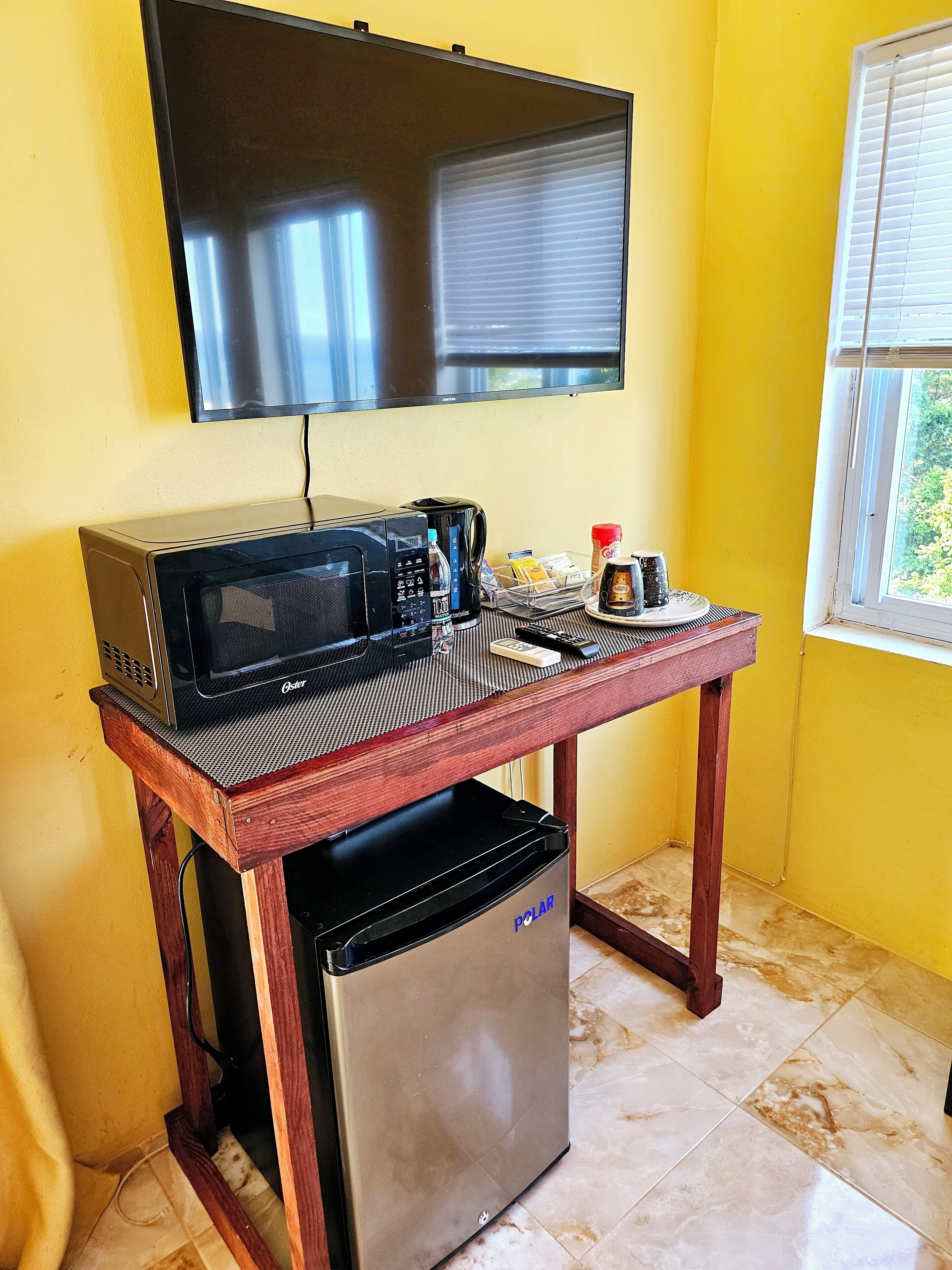 A compact kitchenette with a microwave, toaster, and refrigerator on a wooden table, a television above, and a window with blinds.