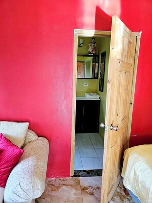 A room with a red wall, a wooden door leading to a bathroom, and a bed with a white and pink pillow. - Boston Bay (Port Antonio)