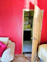 Una habitación con una pared roja, una puerta de madera que da a un baño y una cama con almohada blanca y rosa.