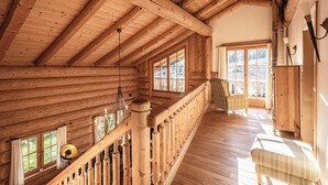 Interior de una cabaña de troncos con una escalera de madera, un banco acolchado y ventanas amplias que dejan entrar luz natural.