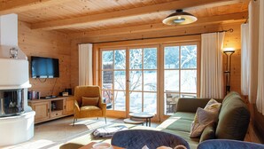Indbydende hyttekøkken med træloft, fjernsyn monteret på væggen, sofa, lænestol, bord med kop og kager, samt pejs.