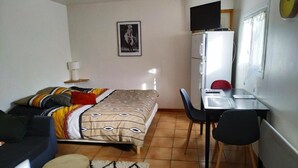 Una habitación pequeña con cama, escritorio, silla, mesita redonda, lámpara y televisor sobre un mueble.