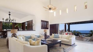 Un salon spacieux avec un grand canapé, une table basse et un lustre. De grandes fenêtres offrent une vue sur l’océan.