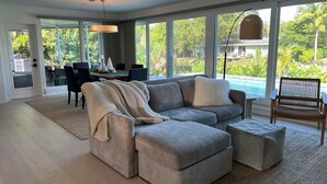 Amplia sala de estar con un sofá seccional gris, un pequeño otoman y un comedor con mesa y sillas. Amplias ventanas ofrecen vistas a una piscina y abundante vegetación.