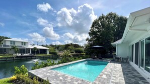 Una piscina con un patio de piedra, vista de una casa y un cuerpo de agua.