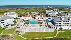 An aerial view of a resort with a central pool, multiple buildings, and a beach in the background.