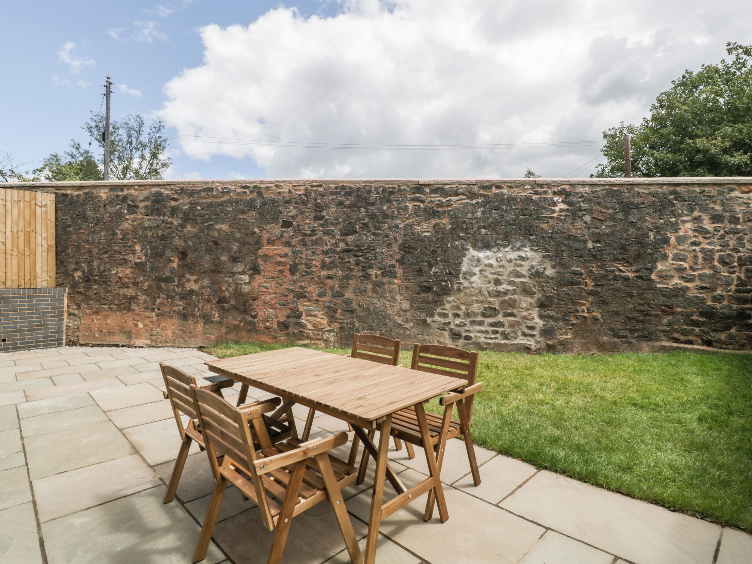 A patio with a wooden table and chairs, a stone wall, and a grassy area.