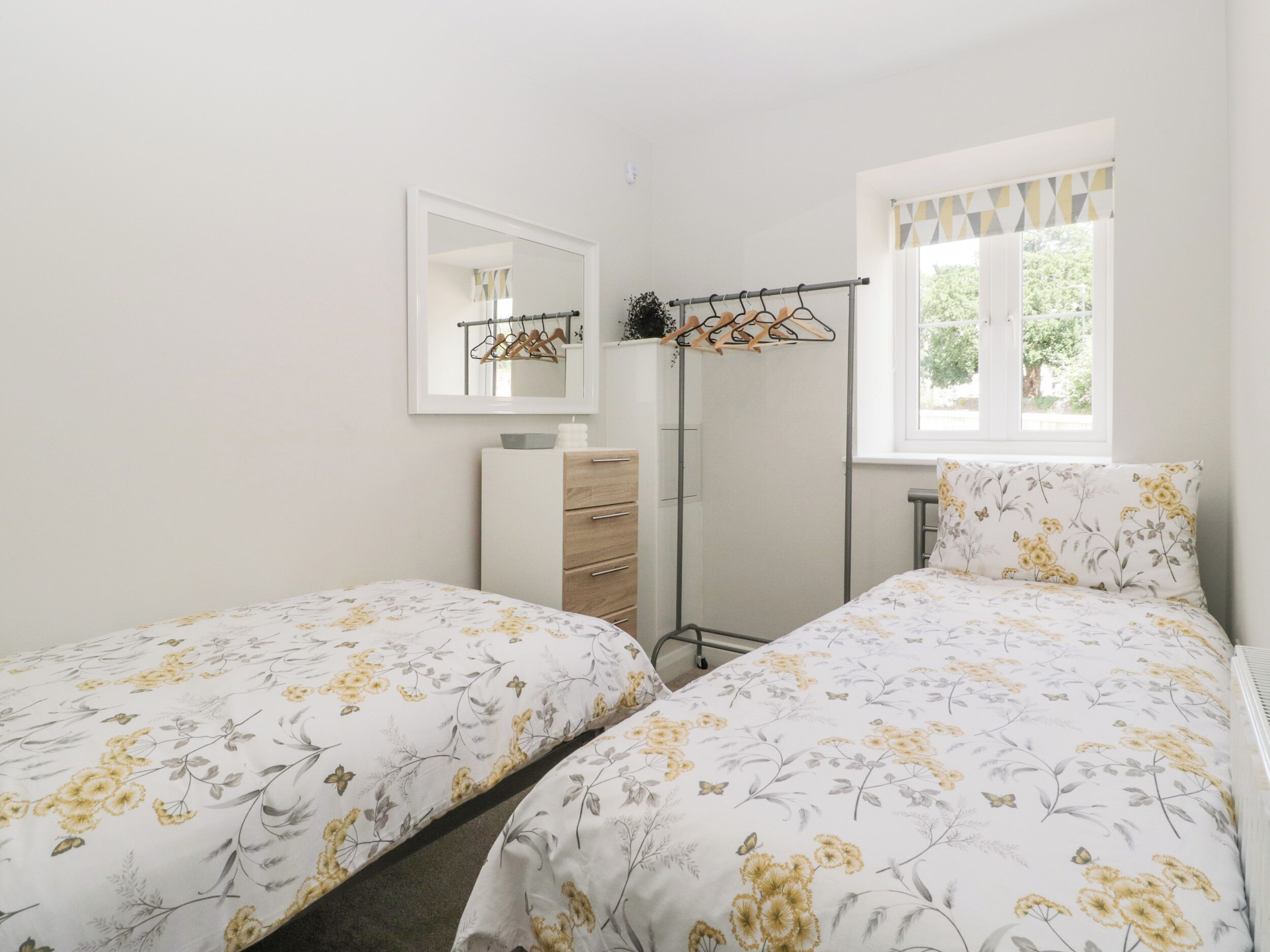 A bedroom with two beds, a dresser, a mirror, a coat rack, and a window with a view of greenery.