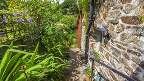 A narrow stone pathway flanked by lush greenery and flowering plants, leading to a rustic stone building with a wooden fence and a metal gate. - Sausage Cottage near Oakford (Nr Bampton)