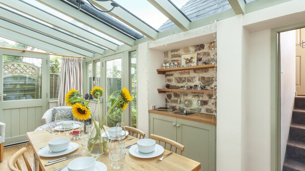 A conservatory dining area with a table set for four, sunflowers, and a stone wall with shelves. - Sausage Cottage near Oakford (Nr Bampton)