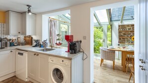 A compact kitchen with a washing machine, a coffee maker, and a dining area with a table and chairs. - Sausage Cottage near Oakford (Nr Bampton)