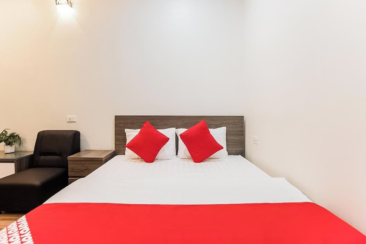 A hotel room with a large bed, two red pillows, a black chair, a small table with a plant, and a wooden headboard.