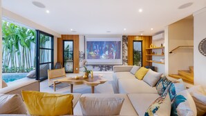 Ein modernes Wohnzimmer mit einer großen Couch, einem Couchtisch und einem Fernseher. Durch die Fenster ist ein Swimmingpool zu sehen.