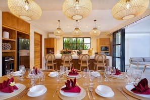 A dining area with a wooden table set for a meal, surrounded by chairs, and a kitchen in the background.