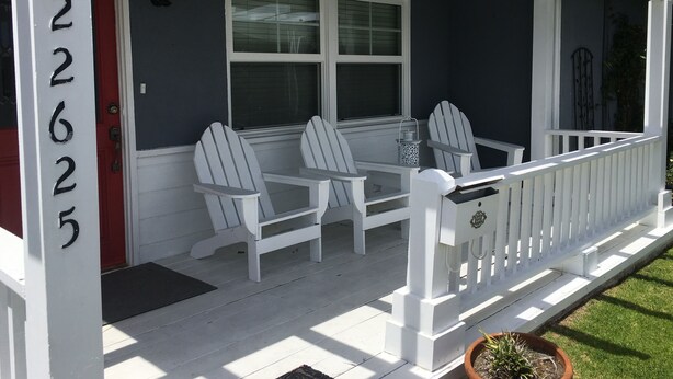 A porch with two Adirondack chairs and a small table, a red door, and a white fence.