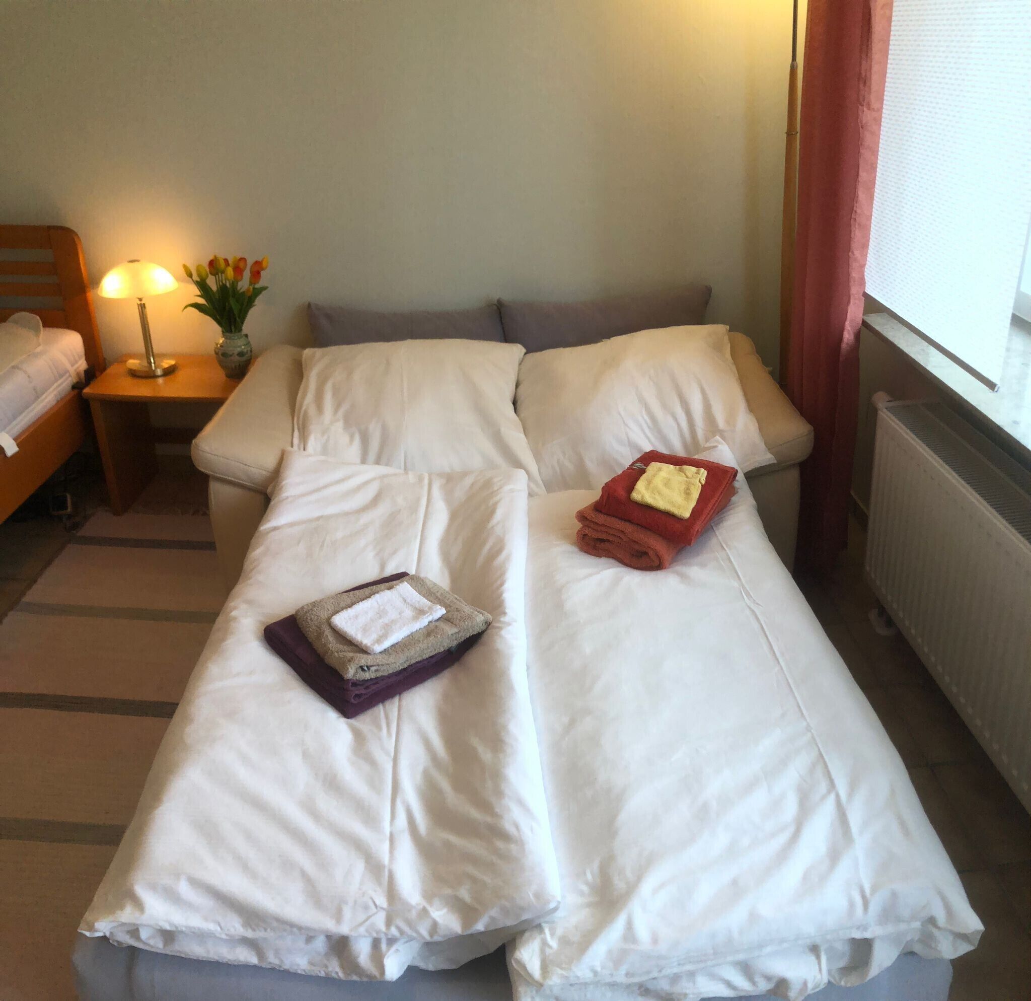 Uma cama de casal com lençóis brancos, uma mesinha de cabeceira com abajur e um vaso com flores.