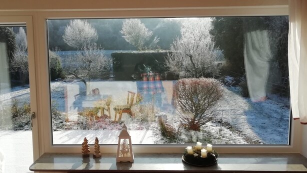 Ein Fenster mit Blick auf einen verschneiten Garten, ein kleiner Tisch mit Kerzen und ein dekorativer Weihnachtsbaum.