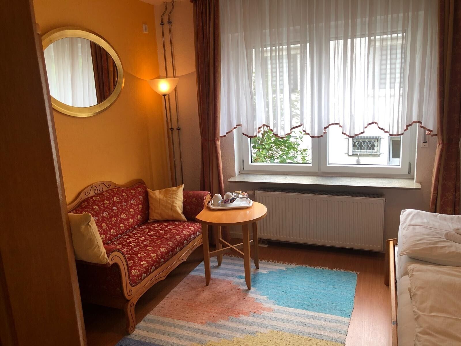 A cozy room with a sofa, a small table with cups and a tray, a window with curtains, and a radiator.