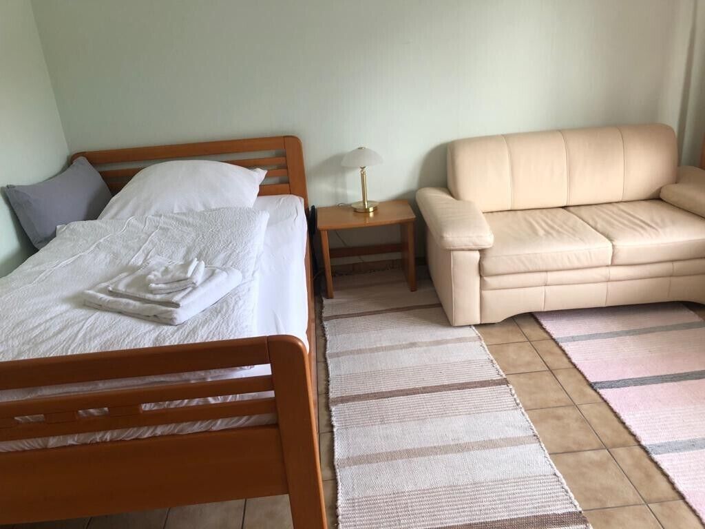 A bedroom with a bed, a bedside table with a lamp, a beige sofa, and a striped rug.