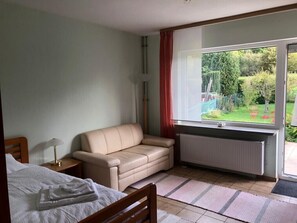 A bedroom with a bed, a sofa, a window with a view of greenery, and a radiator.