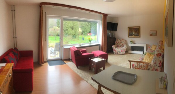 A living room with a red sofa, a red armchair, a floral armchair, a coffee table, and a TV on the wall.
