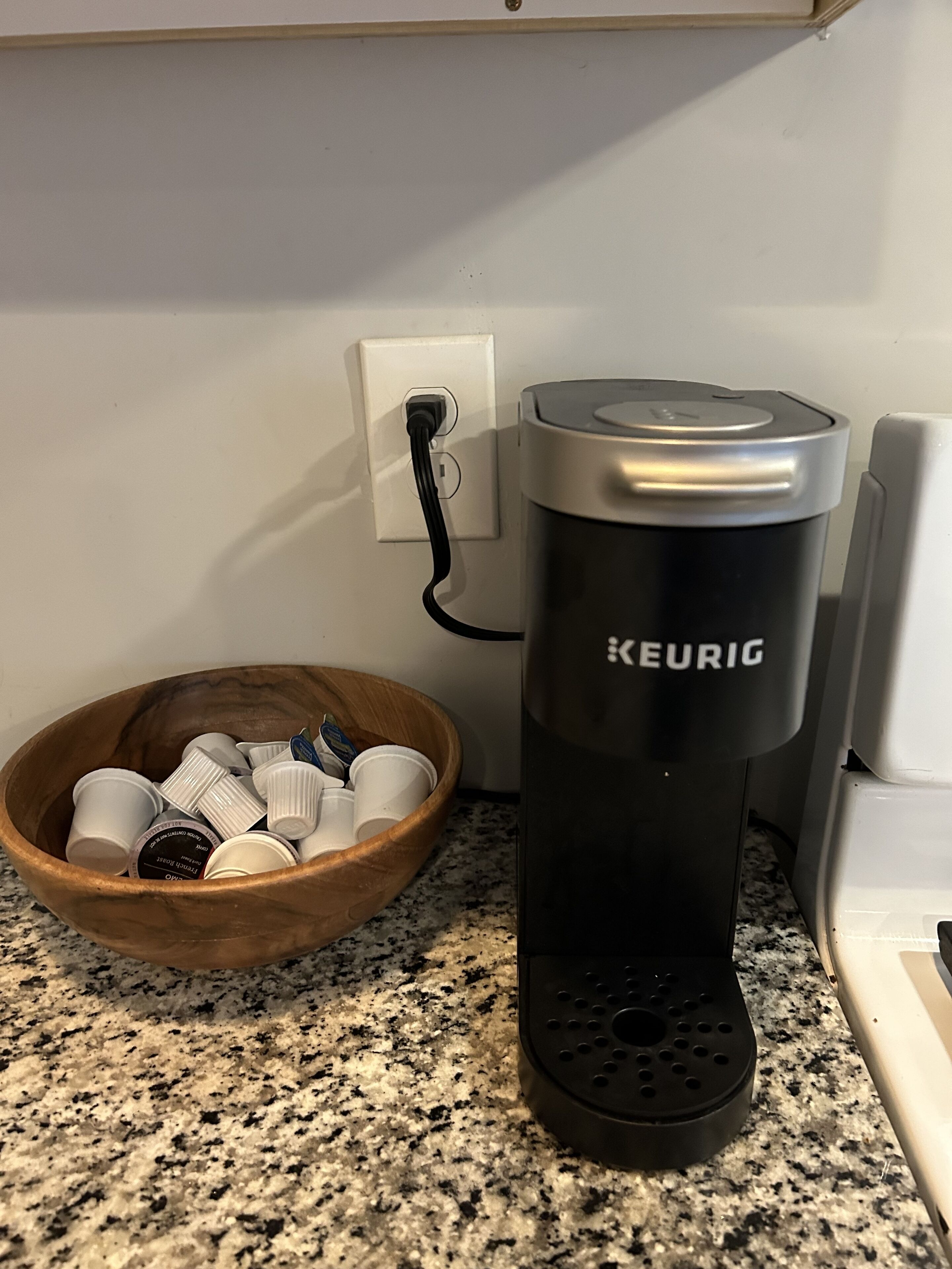 A Keurig coffee maker on a granite countertop, a wooden bowl with coffee cups, and a white electrical outlet.