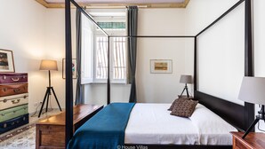 Una cama con dosel, con cubrecama blanco y manta azul, una mesita de noche de madera con una lámpara, un aparador con maletas antiguas y una lámpara de trípode.