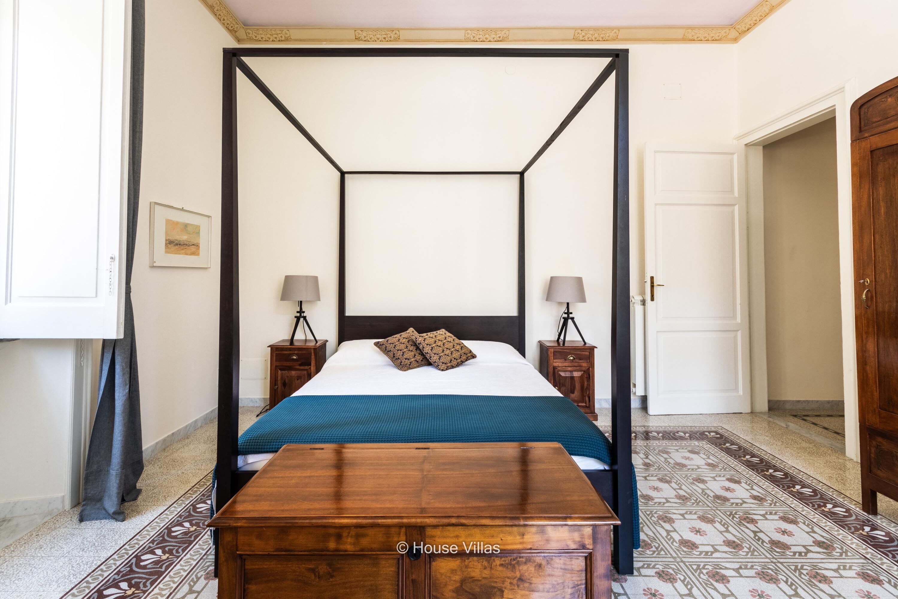 Un lit à baldaquin avec une couvre-lit bleue et blanche, deux tables de chevet avec des lampes, une table de nuit en bois et un sol à motifs.