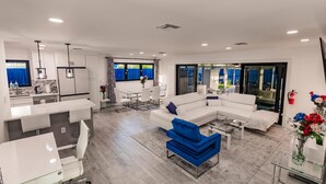 A modern living room with a white sectional sofa, a blue accent chair, and a glass coffee table. There is a kitchen area with white cabinets and pendant lighting. The room has large windows with blue curtains, and a view of the outdoors. - Modern Spacious Home With Pool Jacuzzi (Fort Lauderdale)