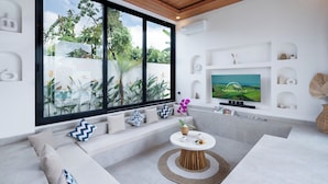 A modern living room with a large TV, comfortable seating, and a view of greenery outside. - Villa Greenland Canggu by Nagisa Bali (Canggu)