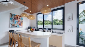 A modern kitchen with a central island, wooden chairs, and a large window with a view of greenery. - Villa Greenland Canggu by Nagisa Bali (Canggu)