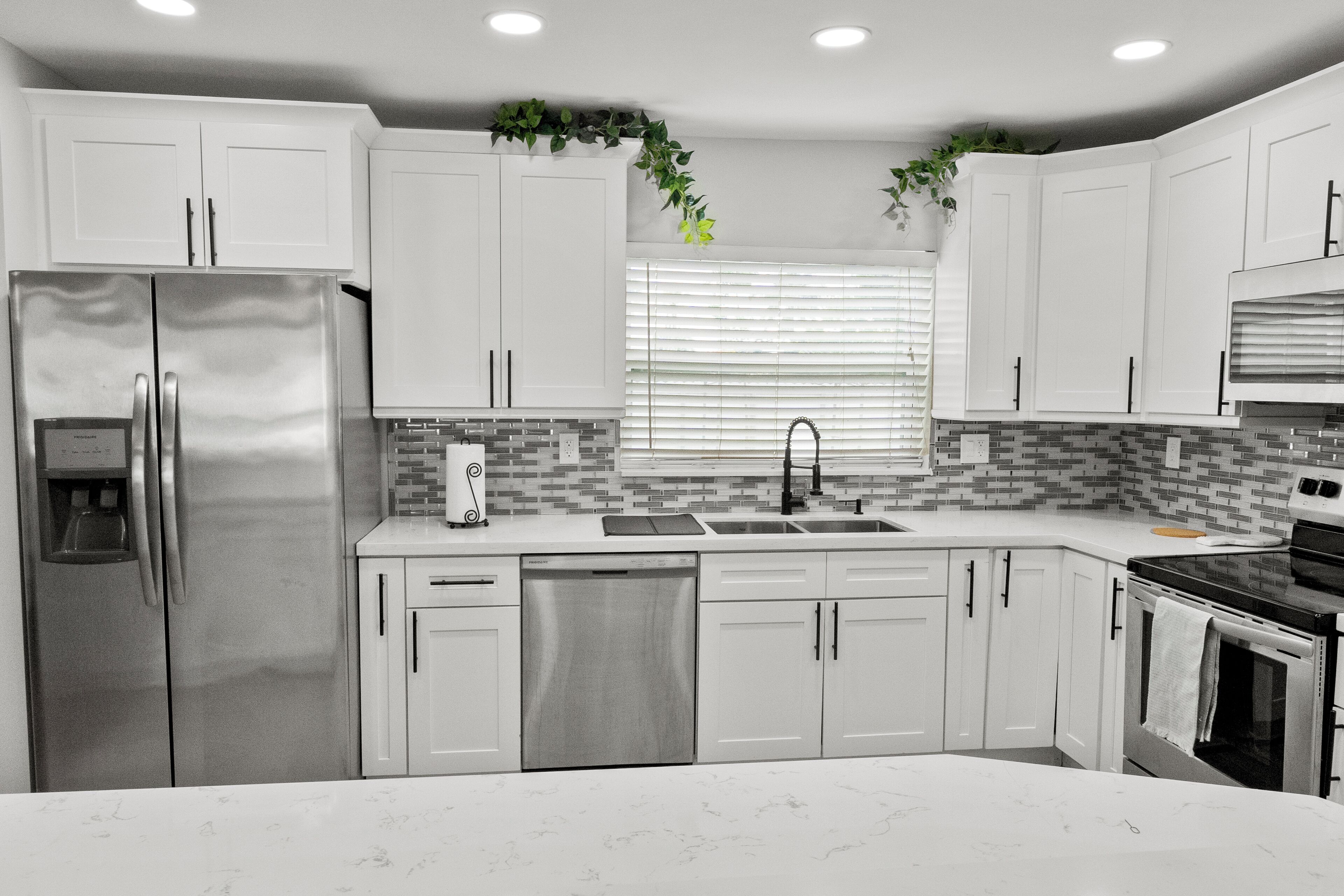 A modern kitchen with white cabinets, stainless steel appliances, a tiled backsplash, and a window with blinds.