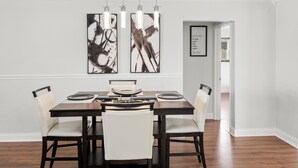 Un comedor con una mesa de madera oscura y sillas blancas tapizadas, dos cuadros abstractos en la pared y un mensaje de bienvenida enmarcado.