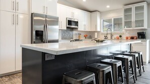 A modern kitchen with white cabinetry, a stainless steel refrigerator, a microwave, and a large island with bar stools. - FoxTrotter | Scottsdale Pool Retreat | Sleeps 12 (Scottsdale)