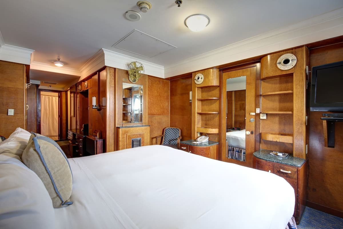 Une chambre d’hôtel avec un grand lit, un bureau avec un téléphone, une chaise, un miroir, une armoire et un téléviseur.