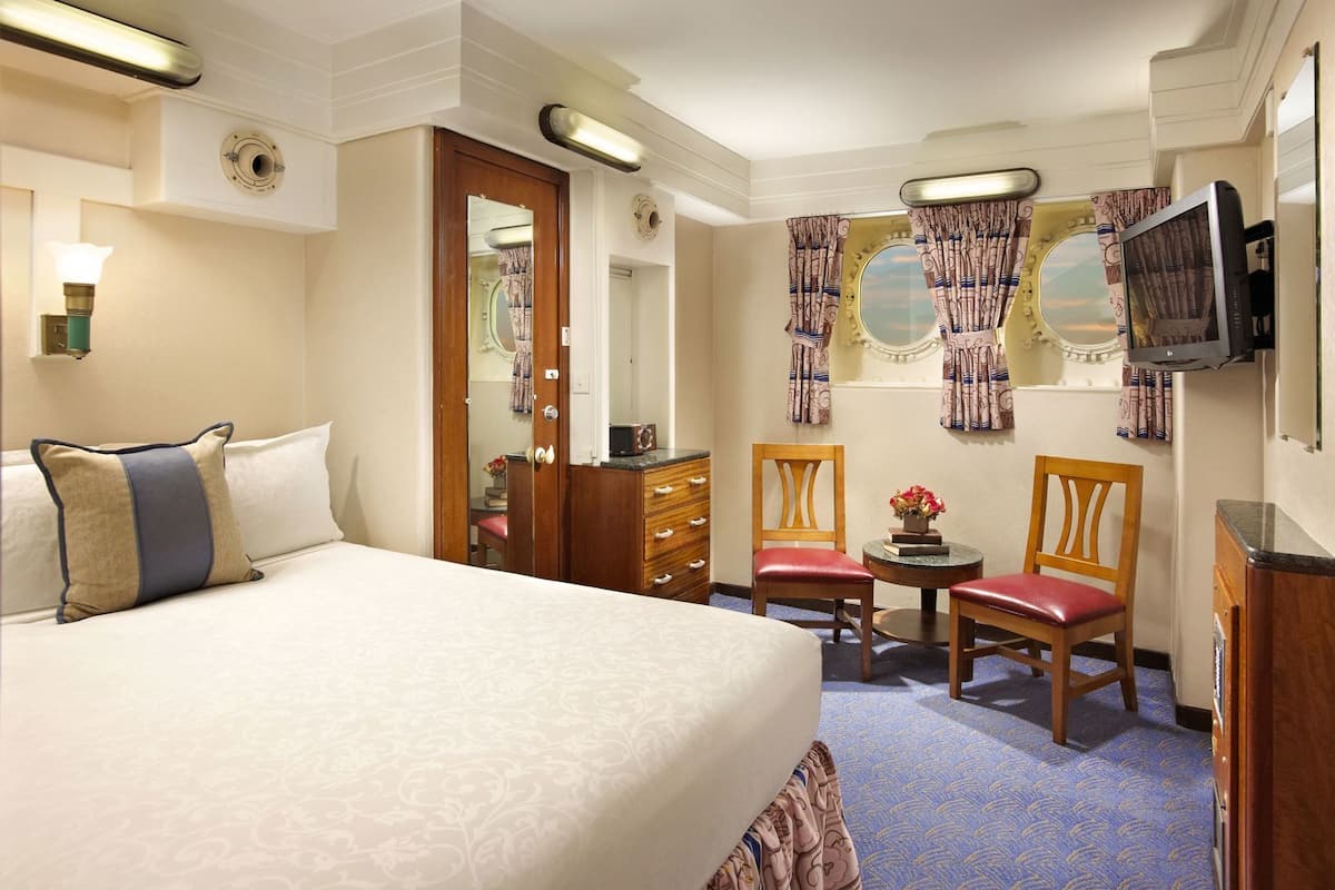 Une chambre d’hôtel avec un grand lit, une commode, deux chaises, une petite table avec des fleurs, un téléviseur et une fenêtre avec des rideaux.