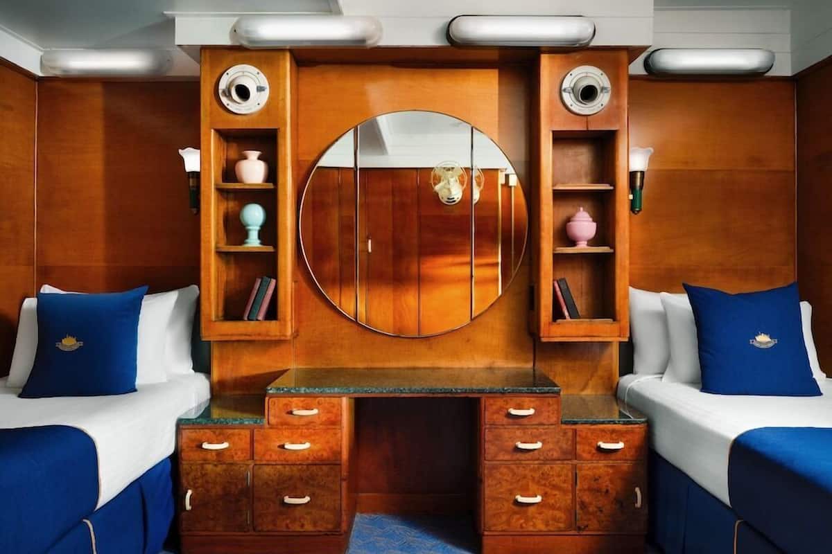 Une chambre avec une coiffeuse en bois, deux lits avec la même literie bleue, et un grand miroir au-dessus de la coiffeuse.