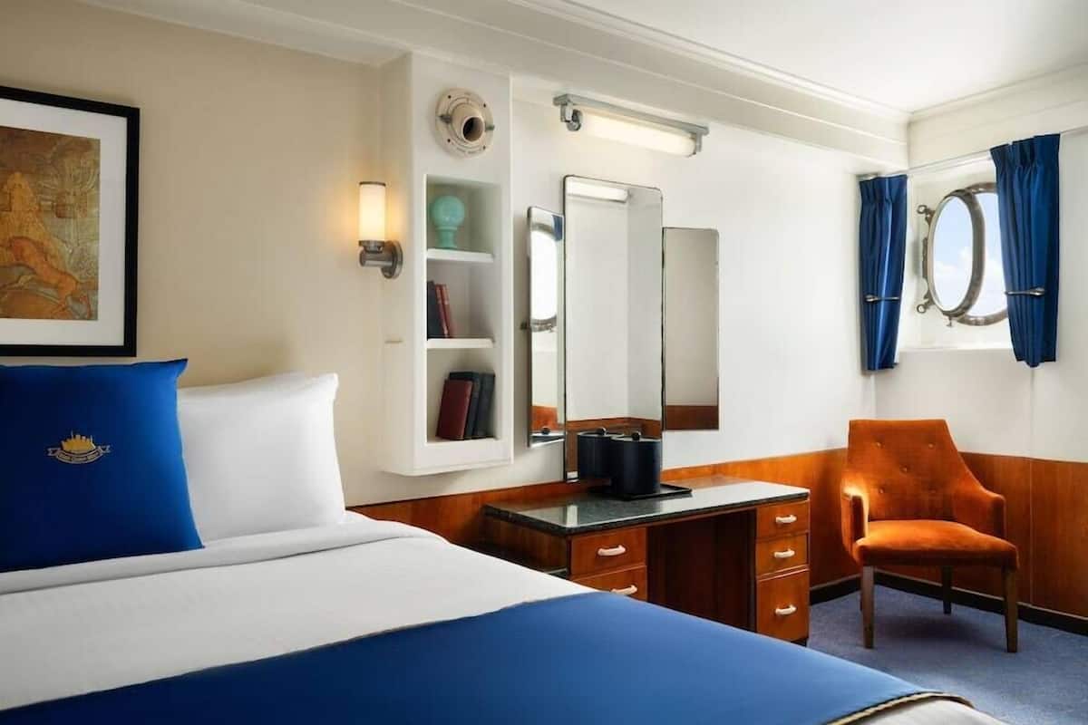 Une chambre d’hôtel comprenant un lit, un bureau, une chaise, un grand miroir, une fenêtre avec des rideaux et une étagère fixée au mur, sur laquelle se trouvent des livres et une lampe.