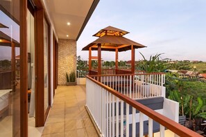 Enjoy the serene views of lush greenery and distant buildings from this balcony with a shaded pergola and white railing.