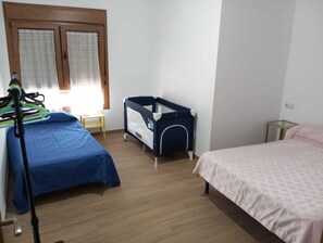 Un dormitorio con dos camas, una cuna, una mesita y una ventana con persianas.